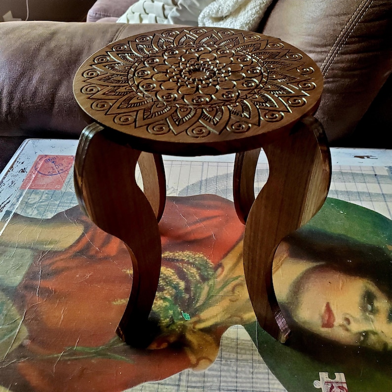 May include: A wooden stool with a round top featuring a carved mandala design. The stool has three legs with a curved shape.