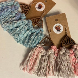 May include: Two pairs of handmade earrings with wooden geometric shapes and colorful tassels. The earrings are made with a light blue and a pink and white color scheme. The wooden shapes are cut in a sunburst design.