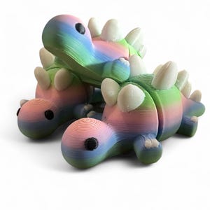 May include: Three small, colorful, 3D printed dinosaur toys. The dinosaurs are stacked on top of each other. The dinosaurs are a light blue, pink, and green color with white accents.