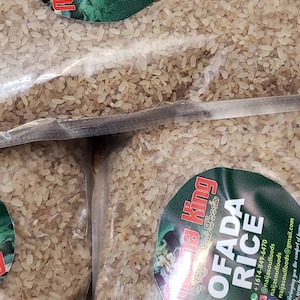 May include: Two clear plastic bags filled with uncooked white rice. The bags have labels with the text "Mama King Naija Soul Foods" and "Ofada Rice" with contact information.