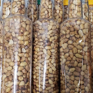 May include: Multiple clear glass bottles filled with roasted peanuts. The bottles are arranged in a row on a shelf.