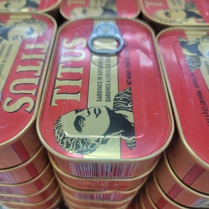 May include: A stack of red and gold canned sardines labeled "TITUS" with the text "SARDINES IN SOYBEAN OIL" and "NET WEIGHT - POIDS NET 125g 4.3 OZ" printed on the label.