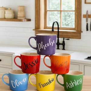 May include: Six brightly coloured ceramic mugs, each with a different name in cursive script. The mugs are stacked and feature the names Oliver, Jessie, Lucy, Frankie, Ashley, and Phoebe. The mugs are blue, red, orange, yellow, green, and purple.