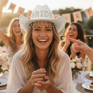 May include: A smiling woman wearing a white cowboy hat with the word "BRIDE" in silver letters. The hat has a beaded fringe. Other women are in the background, celebrating at an outdoor event. The scene is lit by warm sunlight.