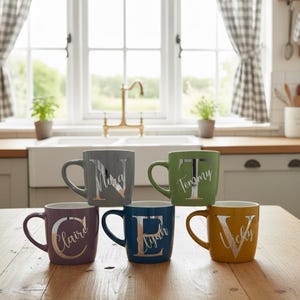 Può includere: Sei tazze in ceramica di vari colori, ognuna con un nome diverso in lettere argentate. Le tazze sono disposte su un tavolo di legno davanti a una finestra. I colori includono grigio, verde, viola, blu e giallo.