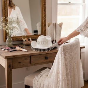 May include: A white cowboy hat with the word "BRIDE" in silver letters. The hat has a beaded fringe and is on a wooden table with makeup and a mirror. A pair of white cowboy boots are in the background.