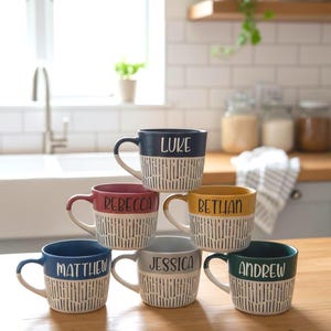 Puede incluir: Una pila de siete tazas de cerámica con nombres impresos. Las tazas tienen una base blanca con un patrón de líneas verticales negras. La mitad superior de cada taza es de un color diferente, incluyendo azul marino, rojo, amarillo, azul, gris y verde.