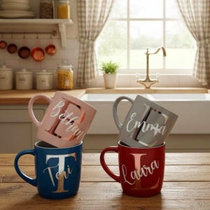Peut inclure: Quatre mugs en céramique roses, grises, bleues et rouges, chacun avec un nom imprimé sur le côté. Les tasses sont empilées sur une surface en bois, avec une cuisine en arrière-plan.