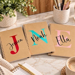 Puede incluir: Tres cuadernos de espiral con cubiertas de color canela. Cada cuaderno presenta una letra mayúscula grande y colorida junto a un nombre escrito en letra cursiva negra. El primero muestra una J roja y Jessica, el segundo una N turquesa y Noah, y el tercero una E rosa y Ella.