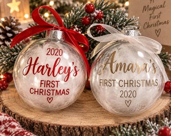 May include: Two clear spherical Christmas baubles filled with white feathers. The left bauble features red text reading Harley's FIRST CHRISTMAS 2020 with a heart and a red ribbon. The right bauble features gold text reading Amara's FIRST CHRISTMAS 2020 with a heart and a white ribbon.