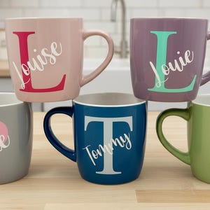 Può includere: Cinque tazze in ceramica di vari colori, ognuna con un nome e un'iniziale diversi. Le tazze sono in tonalità di rosa, viola, grigio, blu e verde. I nomi sono in corsivo, con l'iniziale in grassetto.