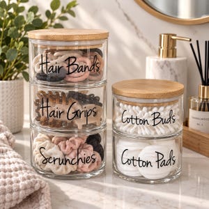 May include: A stack of three clear glass canisters with bamboo lids labelled Hair Bands, Hair Grips, and Scrunchies, alongside a stack of two clear glass canisters with bamboo lids labelled Cotton Buds and Cotton Pads, all on a marble surface.