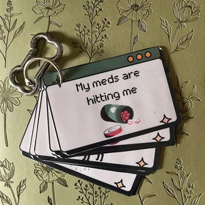 Op de afbeelding: Een set kaarten met de tekst "My meds are hitting me" en een illustratie van een pillenfles. De kaarten zijn bevestigd aan een zilveren sleutelhanger met een botvormig ontwerp. De achtergrond heeft een bloemenpatroon.