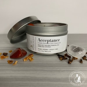 May include: A silver tin candle with the label "Acceptance" and the text "Hand Poured Soy Candles Made with Natural Essential Oils Herbs and Crystals Vegan Organic Cruelty-Free 6 oz | 170 g | Made in USA". The candle is surrounded by various crystals, cloves, and orange peel.