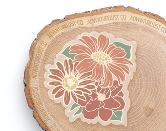 May include: A sticker featuring a bouquet of three flowers in shades of brown and beige. The flowers are stylized and have a vintage aesthetic. The sticker is on a wooden background with the text "Adventurelust Co" repeated around the edge.
