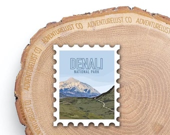 May include: A white stamp with a blue background featuring a mountain landscape and the text "DENALI NATIONAL PARK". The stamp is on a wooden surface with the text "ADVENTURELUST CO" repeated around the edge.