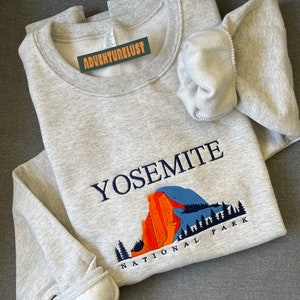 May include: A gray sweatshirt with an embroidered design of Yosemite National Park. The design features a mountain range, trees, and the text "Yosemite National Park".