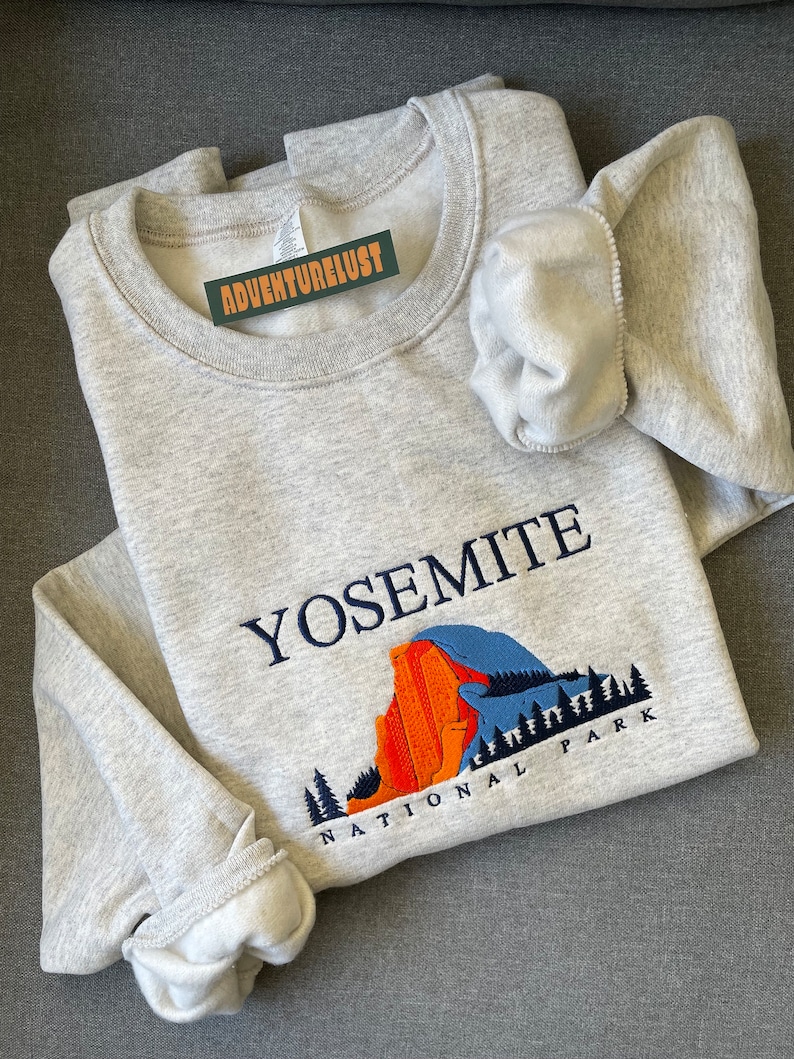 May include: A gray sweatshirt with an embroidered design of Yosemite National Park. The design features a mountain range, trees, and the text "Yosemite National Park".