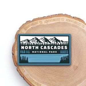 May include: A blue and white sticker with a graphic of mountains and trees. The text "NORTH CASCADES NATIONAL PARK" is printed in black.