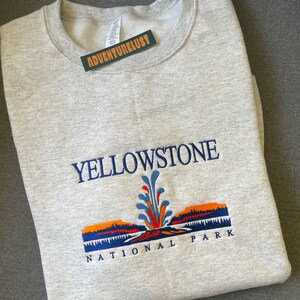May include: Gray sweatshirt with an embroidered design of Yellowstone National Park. The design features a geyser with orange, blue, and green accents, and the text "Yellowstone National Park".