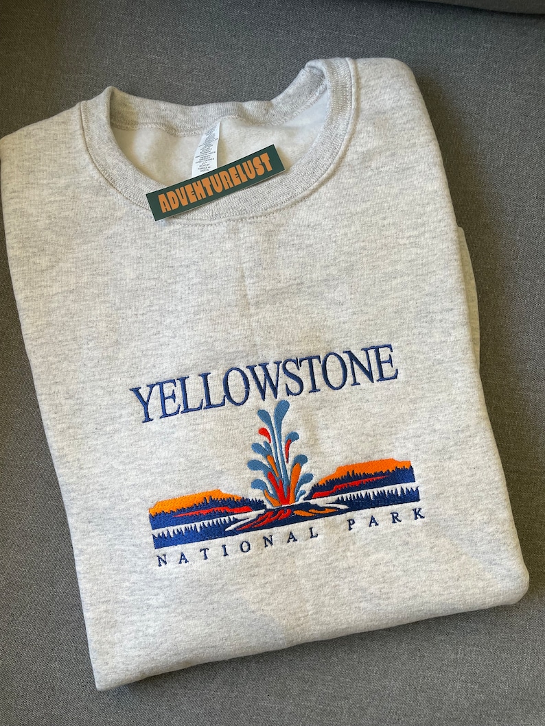 May include: Gray sweatshirt with an embroidered design of Yellowstone National Park. The design features a geyser with orange, blue, and green accents, and the text "Yellowstone National Park".