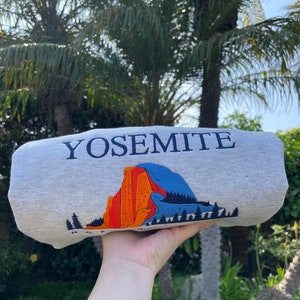 May include: A gray sweatshirt with the word "Yosemite" printed in blue on the front. The sweatshirt features a graphic of a mountain range with orange, red, and blue colors.