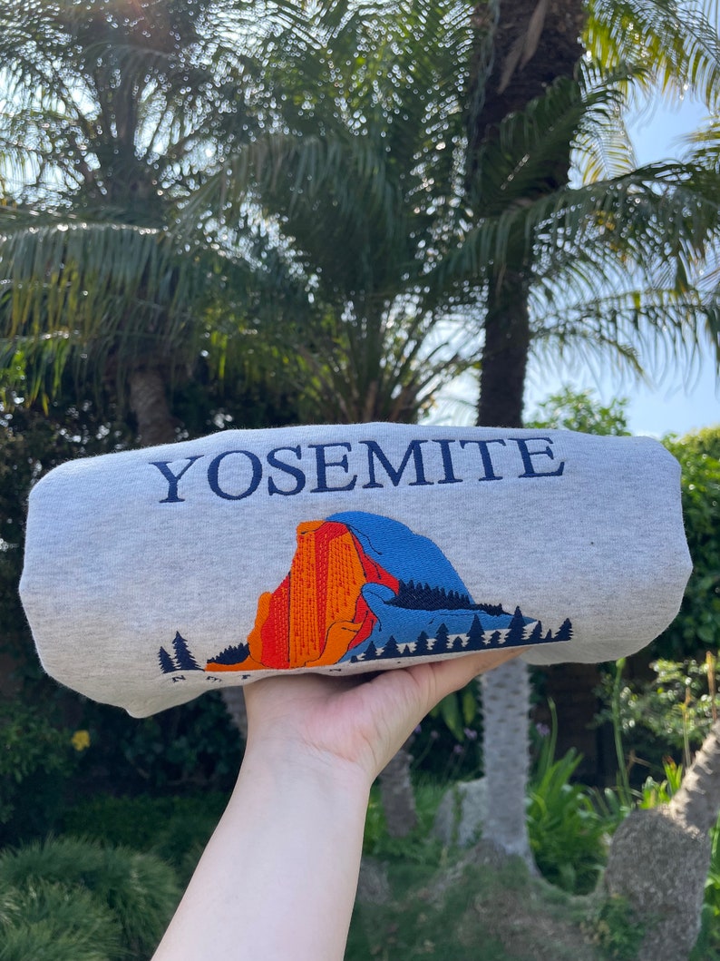 May include: A gray sweatshirt with the word "Yosemite" printed in blue on the front. The sweatshirt features a graphic of a mountain range with orange, red, and blue colors.
