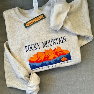 May include: Gray sweatshirt with a graphic of the Rocky Mountain National Park. The graphic features a mountain range with orange peaks and a blue sky. The text "Rocky Mountain National Park" is embroidered in blue on the sweatshirt.