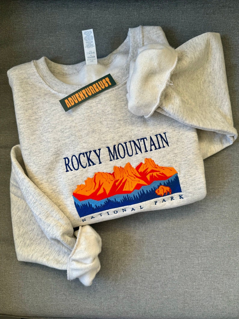 May include: Gray sweatshirt with a graphic of the Rocky Mountain National Park. The graphic features a mountain range with orange peaks and a blue sky. The text "Rocky Mountain National Park" is embroidered in blue on the sweatshirt.