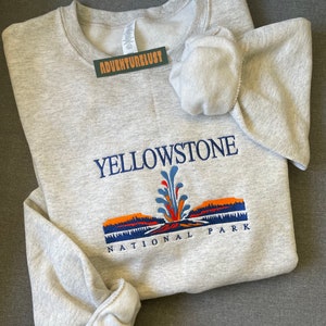 May include: Gray sweatshirt with a Yellowstone National Park logo featuring a geyser and a mountain range in orange, blue, and green colors.