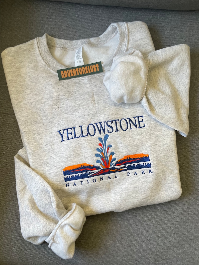 May include: Gray sweatshirt with a Yellowstone National Park logo featuring a geyser and a mountain range in orange, blue, and green colors.