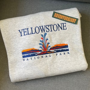 May include: Gray sweatshirt with an embroidered image of Yellowstone National Park. The image features a geyser erupting with a blue, orange, and red color scheme. The text "Yellowstone National Park" is embroidered in blue.