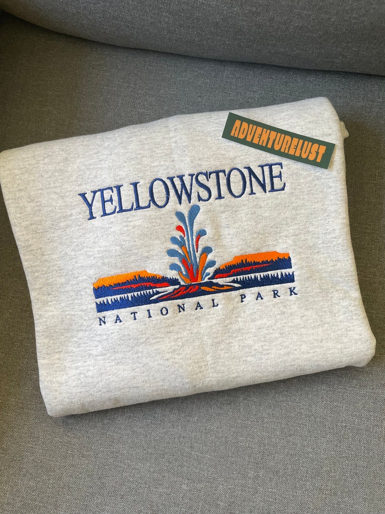 May include: Gray sweatshirt with an embroidered image of Yellowstone National Park. The image features a geyser erupting with a blue, orange, and red color scheme. The text "Yellowstone National Park" is embroidered in blue.