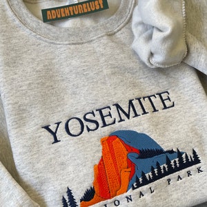 May include: A gray sweatshirt with the words "Yosemite National Park" embroidered on the front. The design features a blue and orange mountain range with trees.