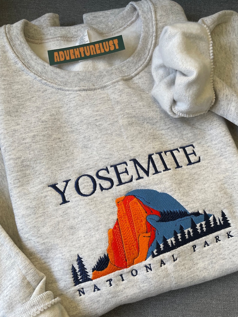 May include: A gray sweatshirt with the words "Yosemite National Park" embroidered on the front. The design features a blue and orange mountain range with trees.