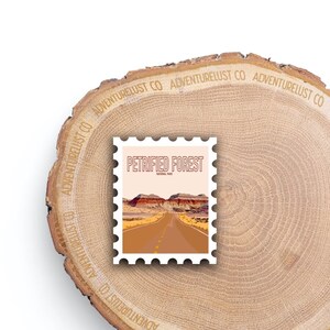 May include: A white sticker with a brown and orange illustration of a road leading to a desert landscape with red rock formations. The sticker says "Petrified Forest National Park" in a red outline.