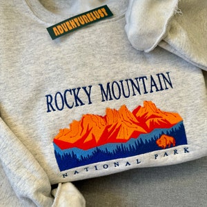 May include: Gray sweatshirt with embroidered text "Rocky Mountain National Park" and a graphic of a mountain range with a bison in the foreground.