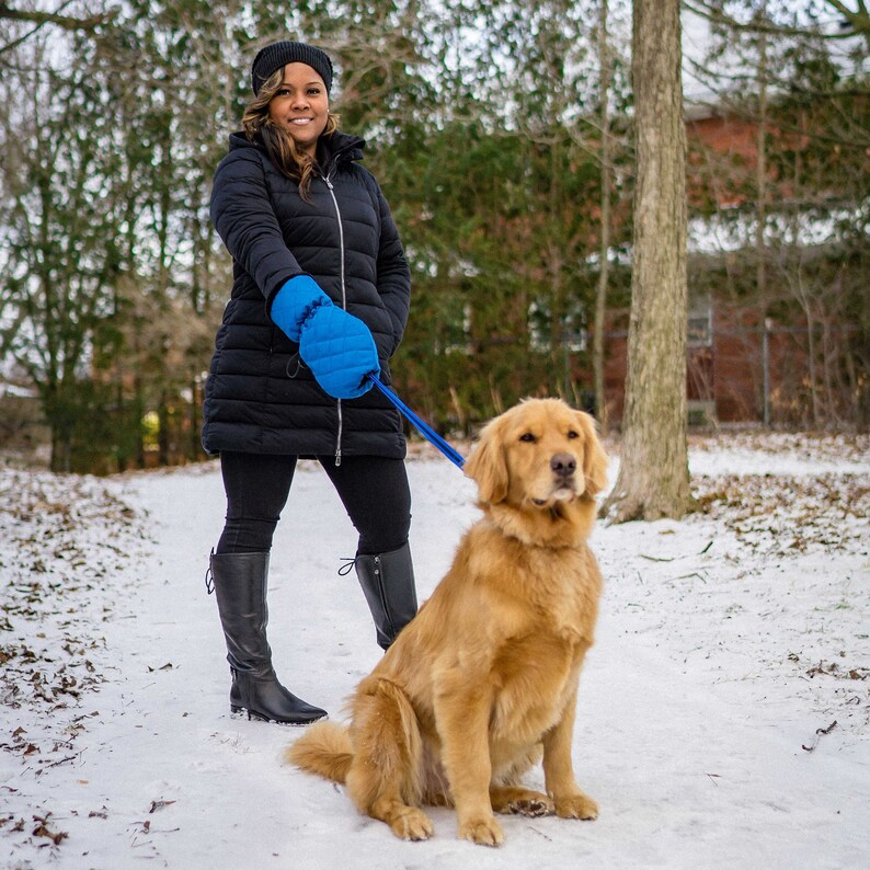 Pode incluir: Uma mulher vestindo um casaco de inverno preto e um chap&eacute;u azul passeia com seu golden retriever na neve. A mulher est&aacute; usando luvas de inverno azuis e o c&atilde;o est&aacute; sentado em um caminho nevado.