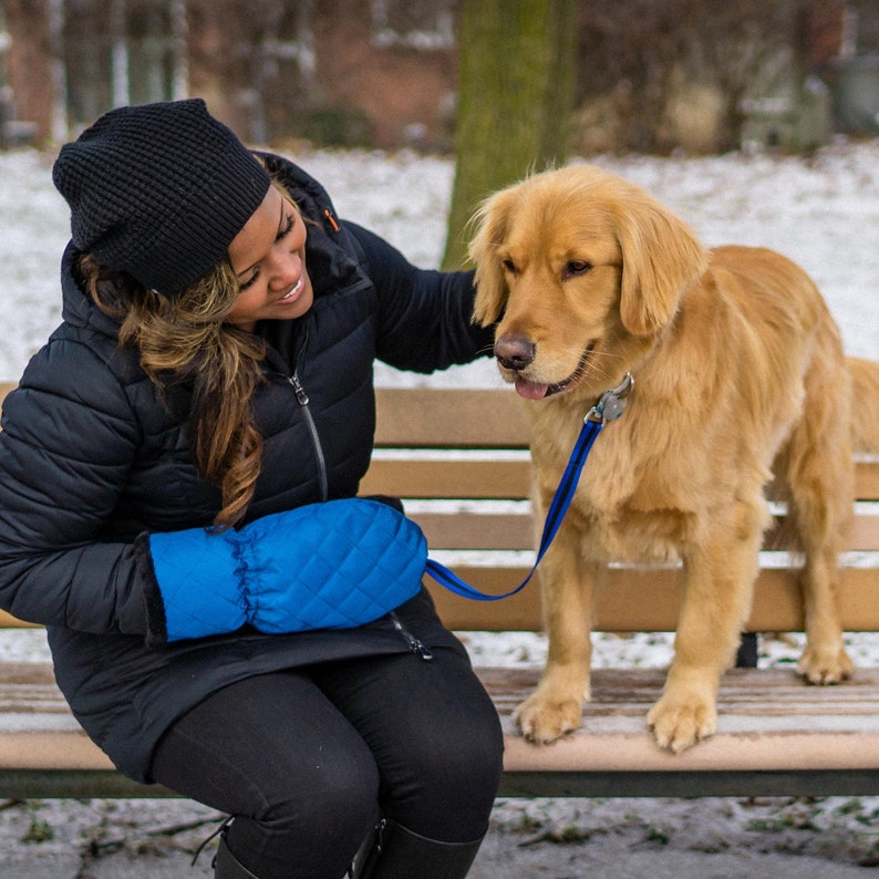 Pode incluir: Uma mulher vestindo uma jaqueta de inverno preta e luvas acolchoadas azuis senta-se em um banco de madeira com um c&atilde;o golden retriever. O c&atilde;o est&aacute; usando uma coleira preta e uma guia.