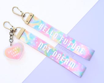 Peut inclure: Deux lanyards colorés avec des porte-clés dorés. Le lanyard du haut porte le texte "HELLO FUTURE" et le lanyard du bas porte le texte "NCT DREAM". Les deux lanyards ont un motif tie-dye avec des couleurs rose, bleu et jaune. Un petit charme en forme de cœur avec le texte "DREAMS COME TRUE" est attaché au lanyard du bas.