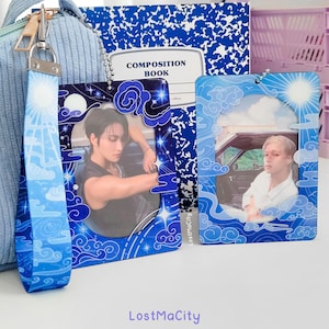 May include: Rectangular keychains with images of people, clouds, and celestial designs, in blue and white. One keychain has a matching blue and white wrist strap. A composition notebook is visible in the background. The keychains have a decorative border.