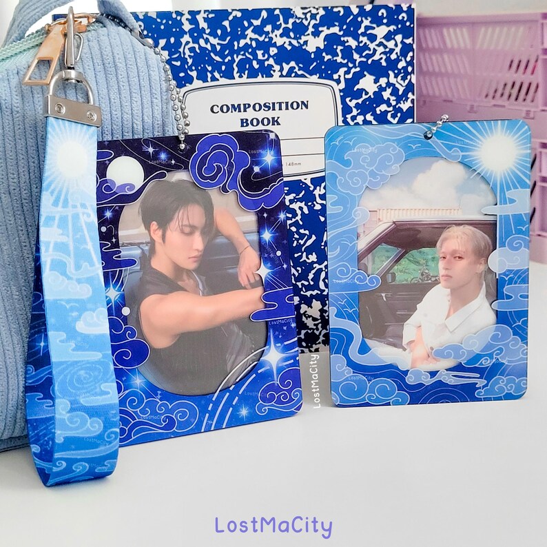 May include: Rectangular keychains with images of people, clouds, and celestial designs, in blue and white. One keychain has a matching blue and white wrist strap. A composition notebook is visible in the background. The keychains have a decorative border.