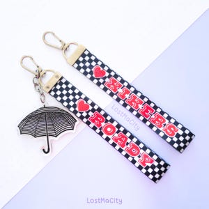 Peut inclure: Deux lanières de porte-clés à carreaux noirs et blancs avec des cœurs rouges et les mots "Road Trip" et "Hikers" imprimés dessus. Un charme de parapluie en acrylique transparent est attaché à l'une des lanières.