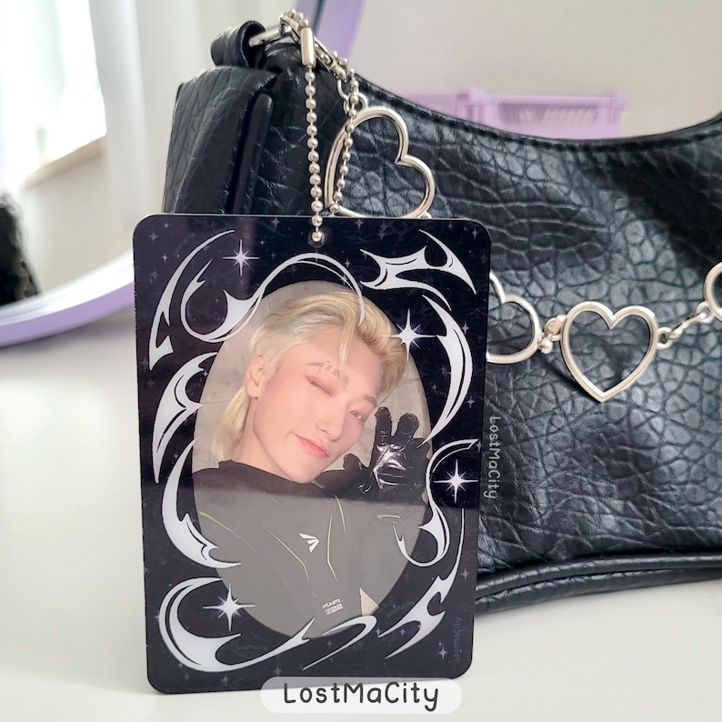 Puede incluir: Un llavero rectangular negro con una foto de una persona con cabello rubio haciendo el signo de la paz. El llavero tiene un dise&ntilde;o decorativo blanco y el texto "LostMaCity". Un bolso negro con una cadena en forma de coraz&oacute;n est&aacute; en el fondo.