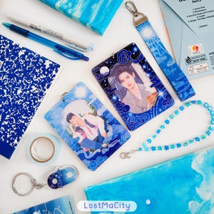May include: A collection of blue and white stationery and accessories, including pens, notebooks, and a beaded lanyard. The image features photo cards with images of a person, a keychain, and a lanyard with a sun and cloud design. The text "LostMaCity" is visible.