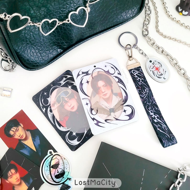 Puede incluir: La imagen muestra una colecci&oacute;n de productos. Se ven dos tarjetas rectangulares con retratos y bordes decorativos. Tambi&eacute;n hay un llavero con una correa estampada y un peque&ntilde;o dije. Una bolsa verde oscuro y cadenas plateadas est&aacute;n en el fondo. El texto "LostMaCity" es visible.