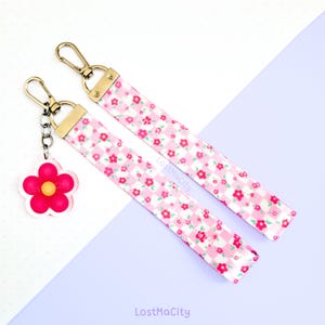 May include: Pink and white gingham keychain with a pink flower charm. The keychain has a pink and white gingham pattern with small pink flowers. The charm is a pink flower with a yellow center. The keychain is attached to a gold chain and a gold clasp. LostMaCity is printed on the keychain.