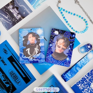 May include: A selection of blue and white merchandise, including photo cards, a bookmark, and a beaded phone strap. The items feature images of people and have a celestial theme with clouds and stars. The text "LostMaCity" is visible.