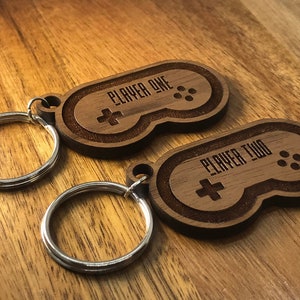 May include: Two wooden keychains shaped like video game controllers. The top keychain says "Player One" and the bottom keychain says "Player Two".
