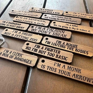 May include: A collection of rectangular wooden keychains, each featuring a small hole and laser-engraved text related to fantasy character classes. Phrases include I'm a Ranger, I'm a Sorcerer, I'm a Warlock, I'm a Wizard, I'm an Artificer, I'm a Bard, I'm a Barbarian, I'm a Paladin, I'm a Druid, I'm a Rogue, I'm a Fighter, I'm a Cleric, and I'm a Monk, along with humorous class-specific commentary.